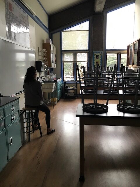 A dark image of a high school classroom that is empty except the teacher.