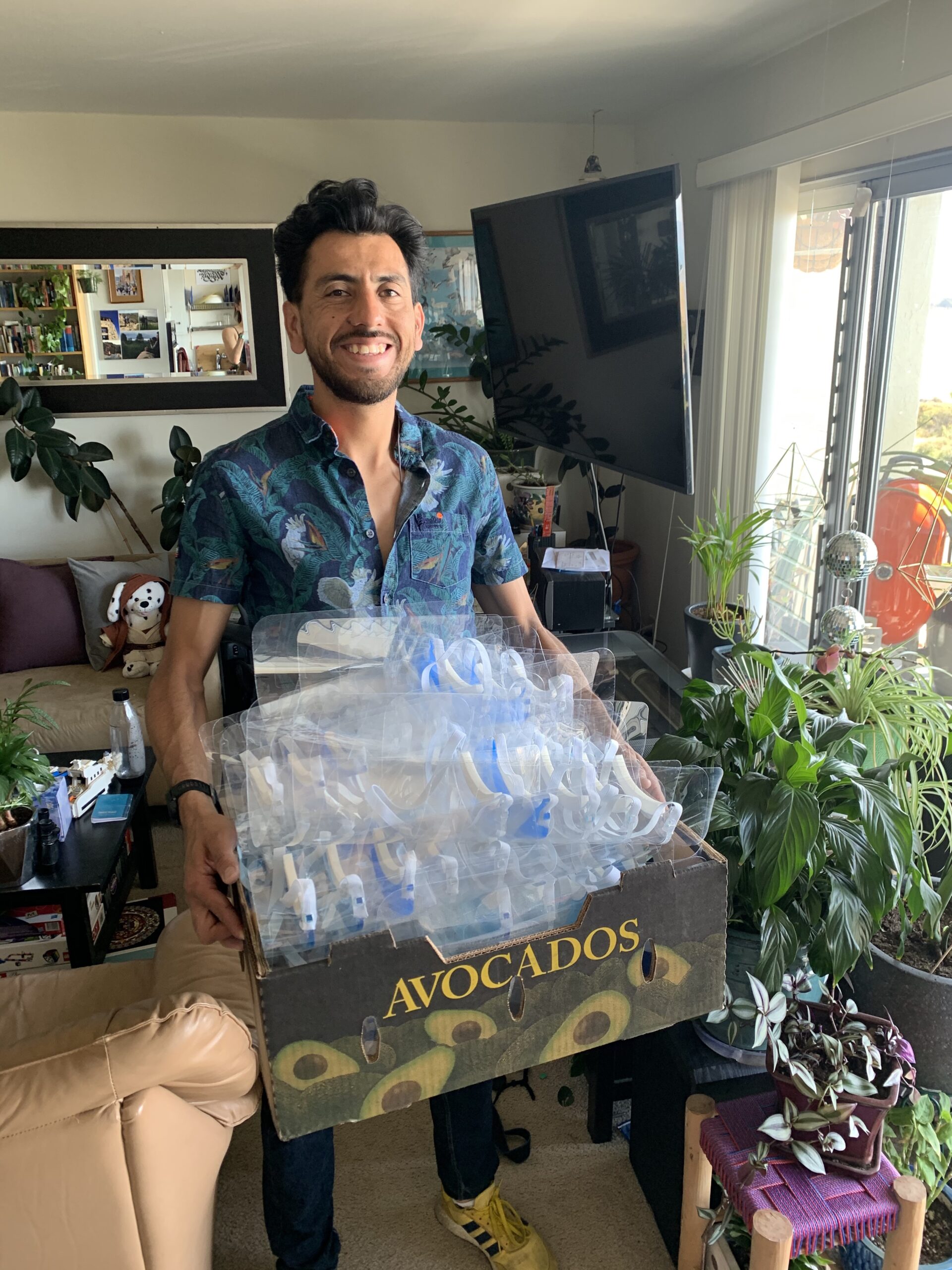 A smiling man carries a cardboard box filled with plastic face shields for Kaiser nurses. The cardboard box says "avocados" on it.