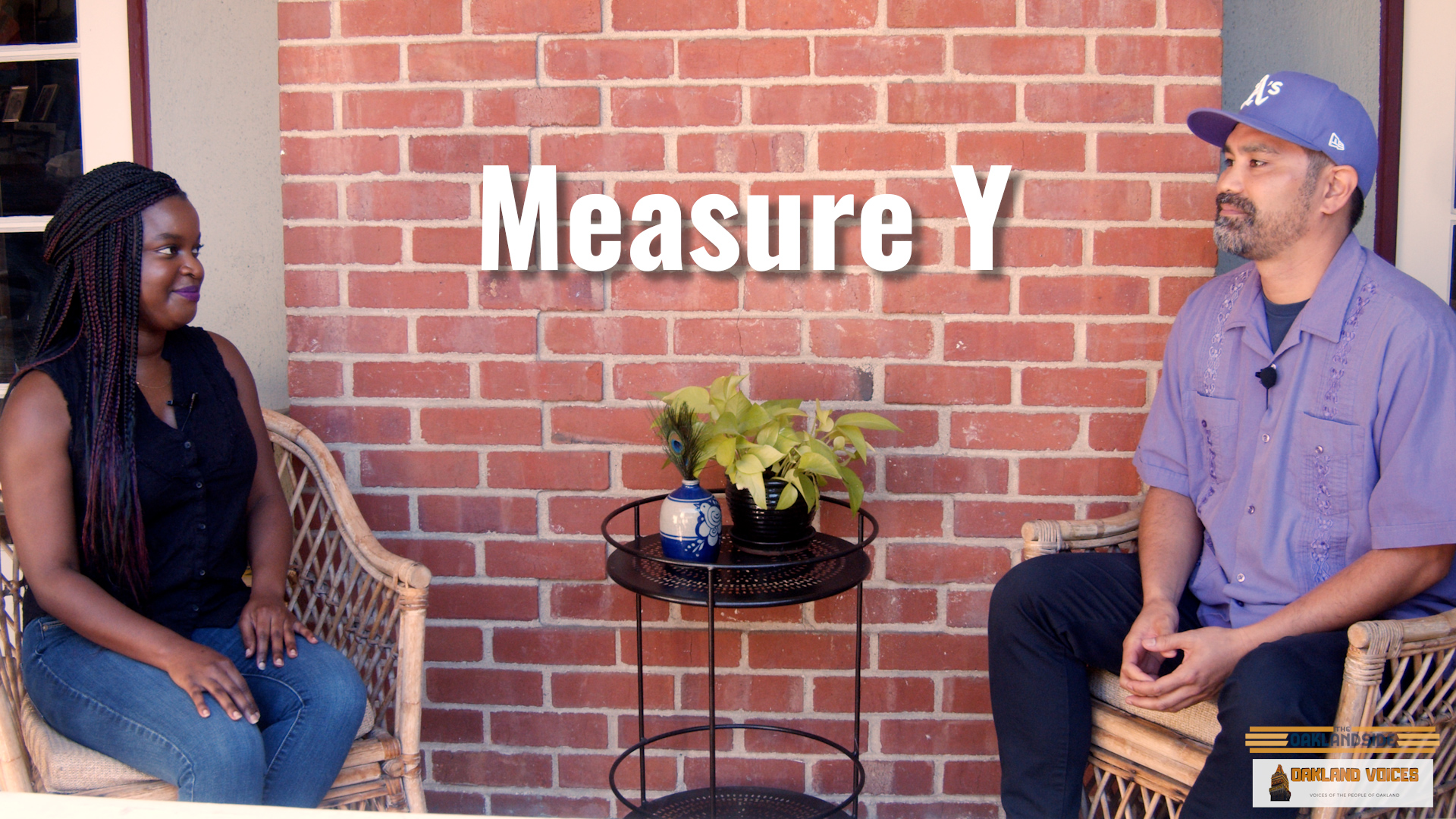 An African American woman with braids sits on a porch chatting with a Filipino man wearing a purple hat and shirt.
