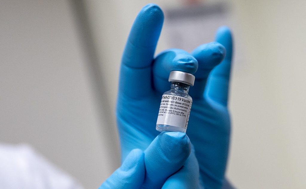 An image of a hand in blue gloves holding a vial of the COVID-19 vaccine.