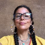 A Mexican American woman wearing two long braids, hoop earrings and glasses and bright embroidered yellow shirt