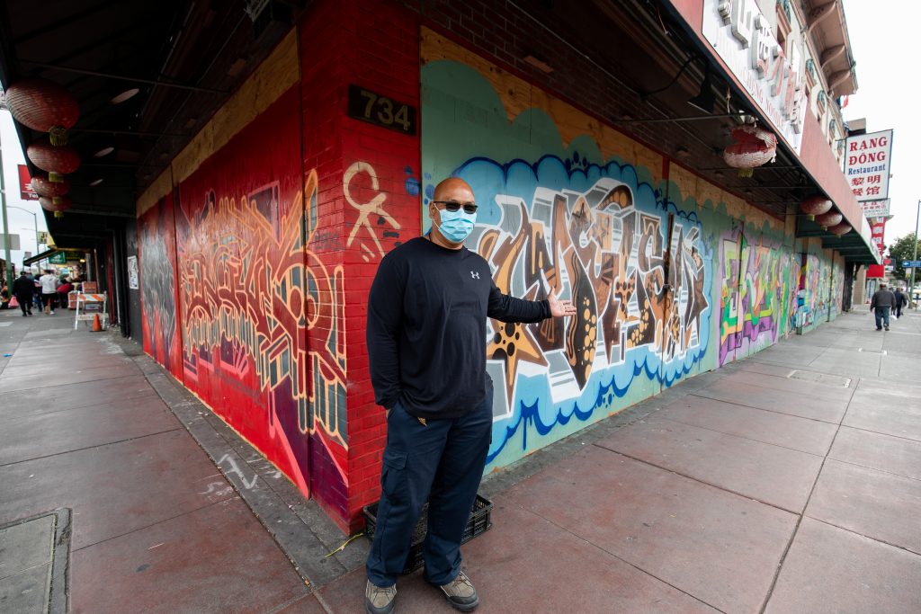 A tall, bald Lao and Chinese man wearing a facemark stands at a corner in Chinatown.