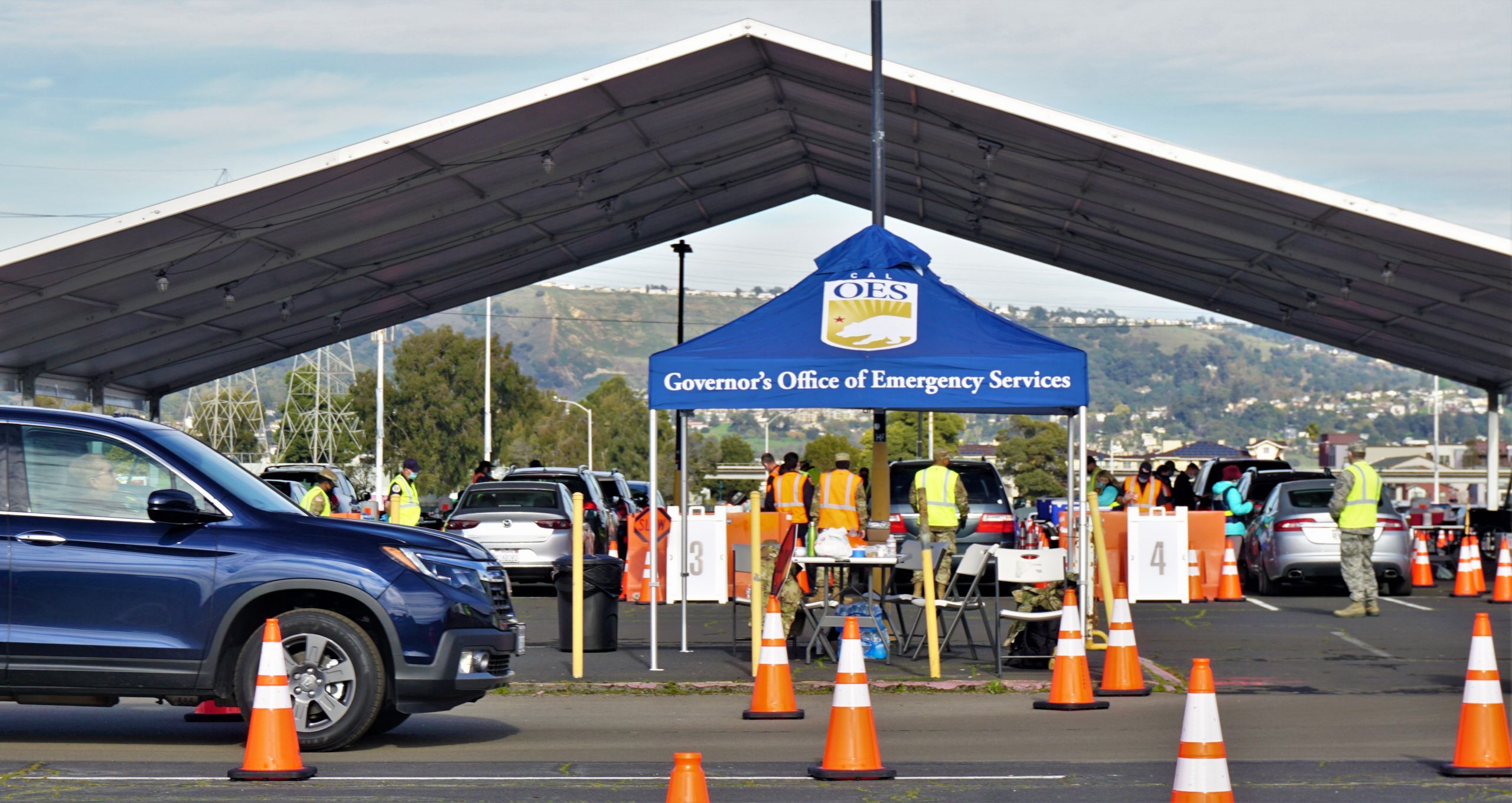 Orange cones line the way in a parking lot at the Oakland Coliseum, where military dressed people point the way. A big blue tent that says "Governor's Office of Emergency Services" is set up.