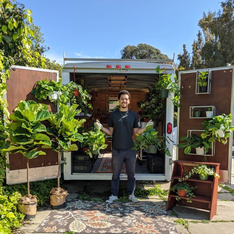 An African American/Filipino man stands in front of a trailer opened in the back that is filled with plants.