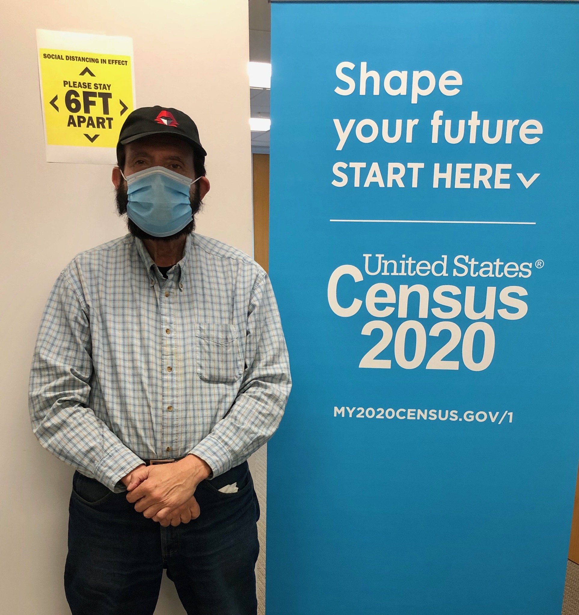 A 74 year-old man wearing a baseball cap and baby blue surgical mask stands in front of sign that says Census 2020 and :"six feet apart"