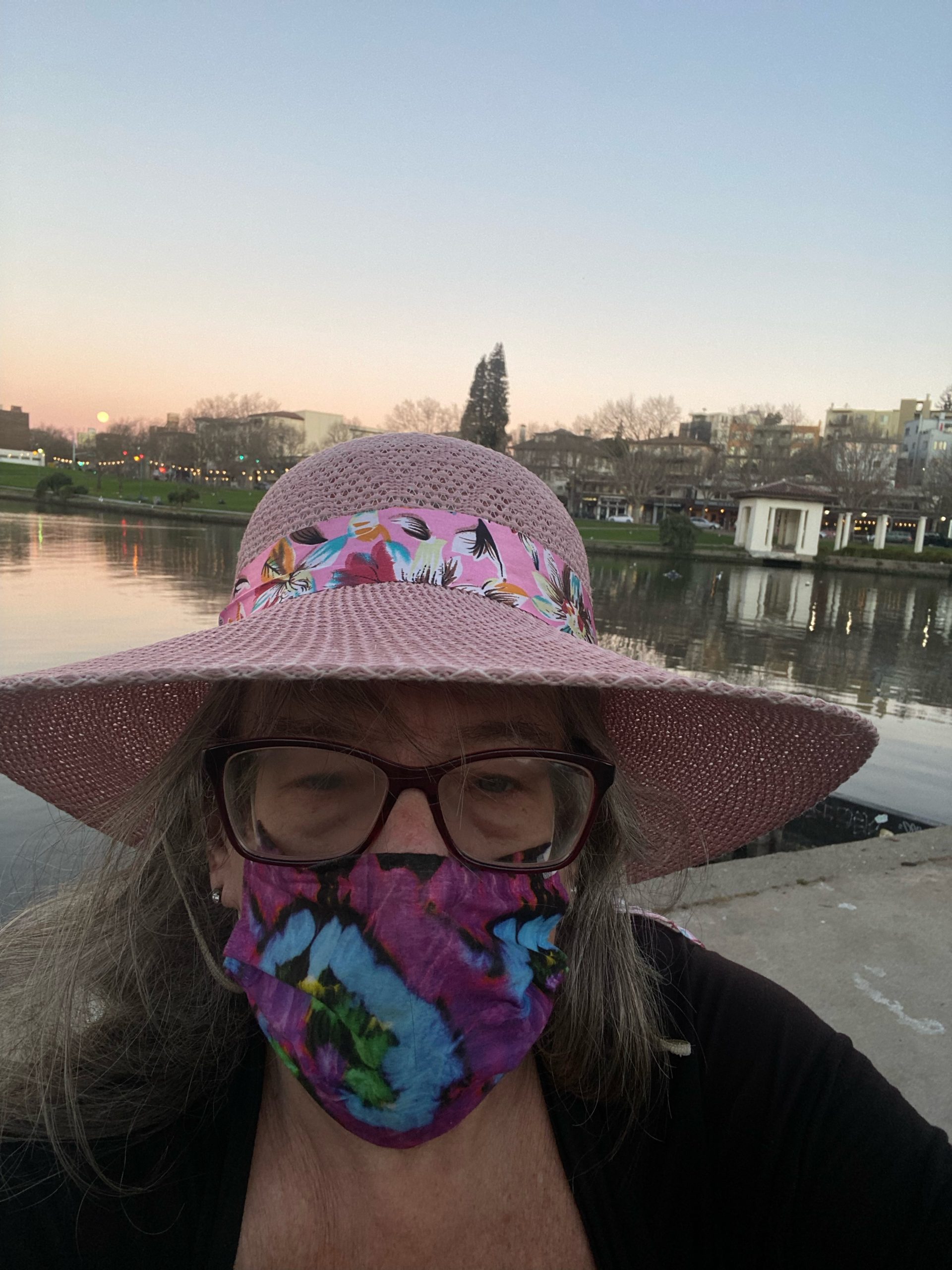 A woman wearing a purple mask, pink hat, and glasses takes a selfie in front of Lake Merritt.