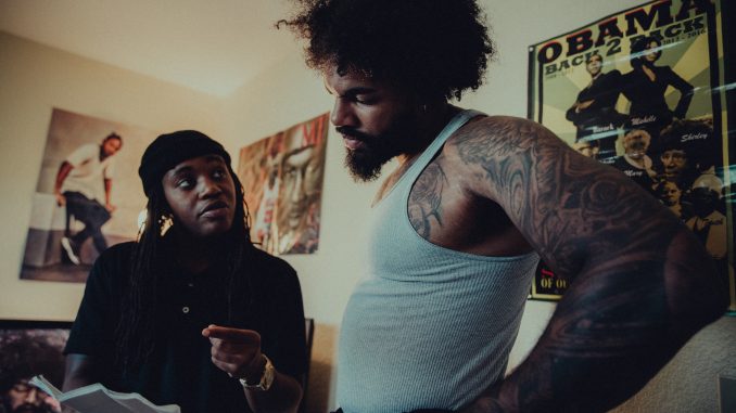 An African American director with a beanie chats with a tall man in a white tank top and with an Afro