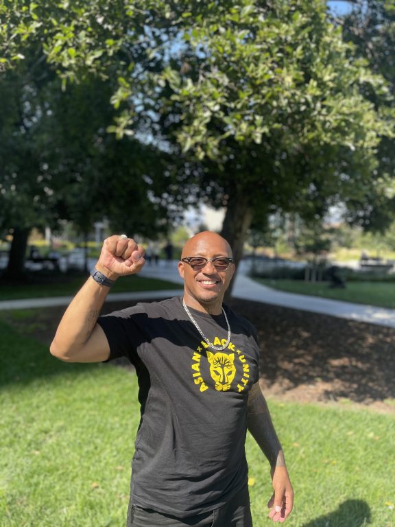 Black man with sun glasses and bald head wearing black shirt with yellow "Black Asian Unity" text stands with fist raised at Lake Merritt