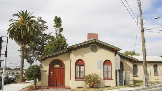 A beige and brick colored small quaint church next to palm tree and electric lines, with gold painted fan details on the doorway and a gold cross on the front of church