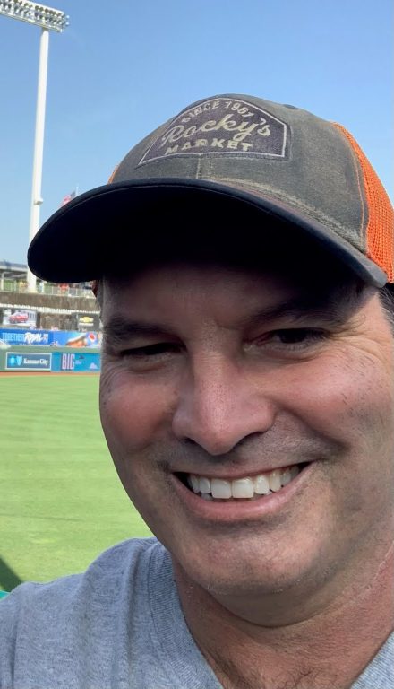 A man wearing baseball cap smiles with teeth at baseball game