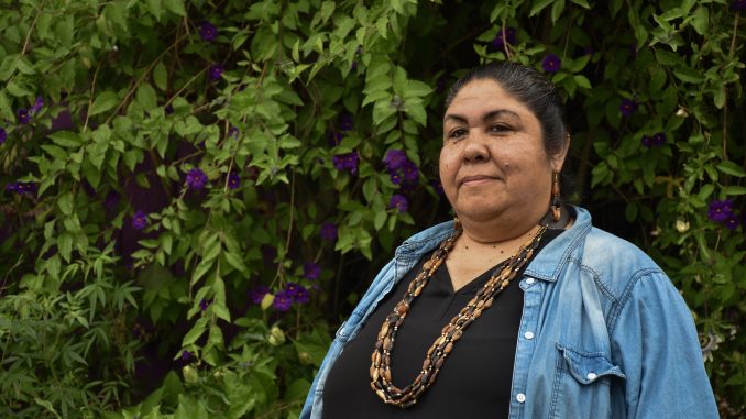 An Indigenous woman wearing denim and beaded necklace stands in front of green trees