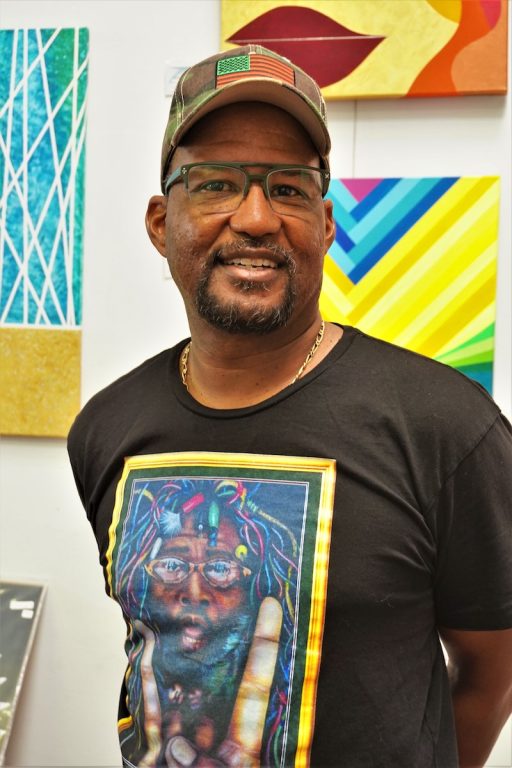 An African American man wearing glasses and baseball cap smiles for camera with colorful paintings in background