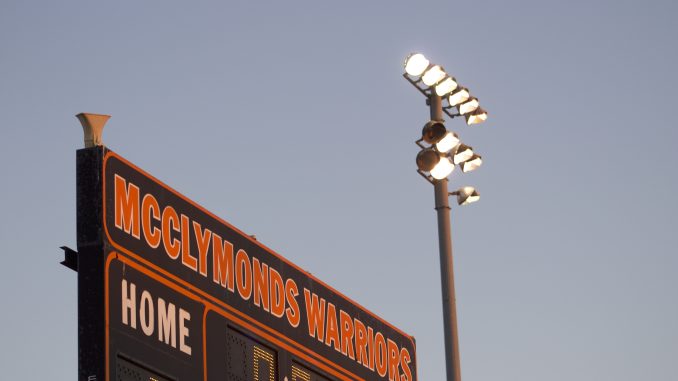 a close up of a scoreboard that says "McClymonds Warriors" in orange type. A field lamp post is shining at dusk next to it