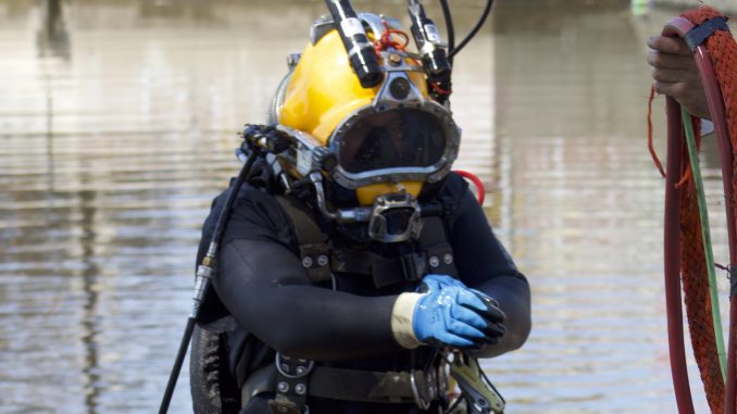 A man in a wetsuit and a yellow hard helmet comes out of murky waters
