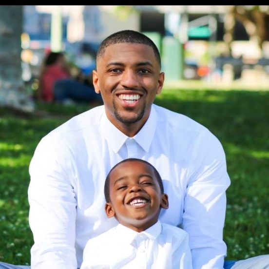 An African American man wearing a white collared shirt hugs a young African American boy in an outdoor portrait