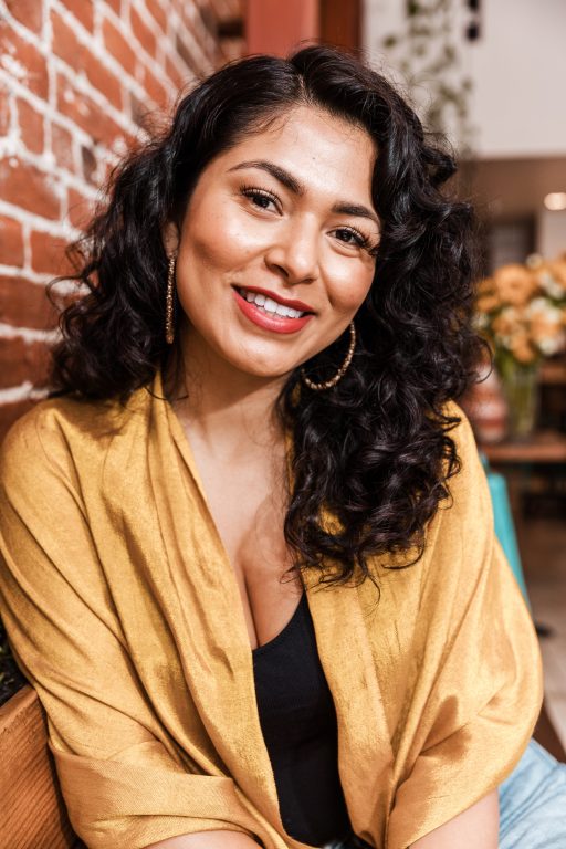 A Latina woman with curly brown hair smiles for camera inside a restaurant with brick wall