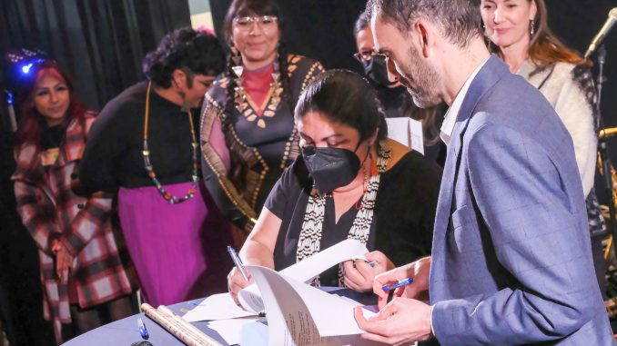 An Ohlone Indigenous woman with long hair wearing a face mask signs and agreement at a public event