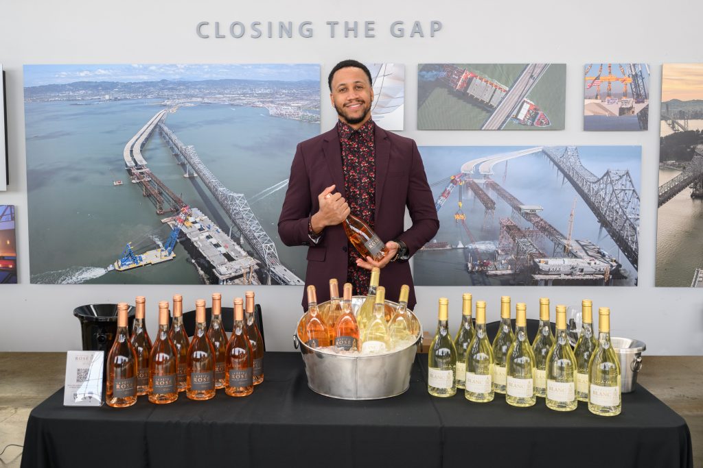 A mixed race African American man wearing a nice Burgundy suit holds up a bottle of rose behind a table showcasing many wines