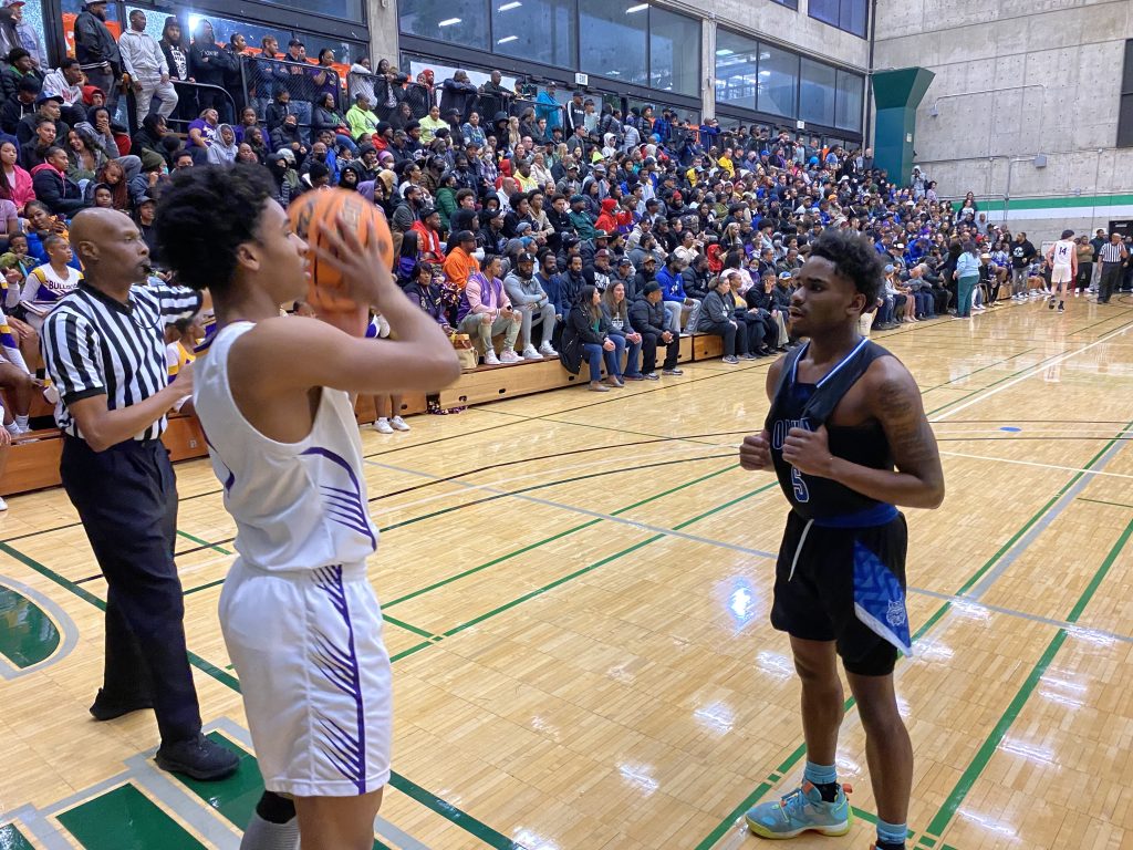 An African American teen wearing a white jersey inbounds ball while an African American teen wearing black jersey guards him