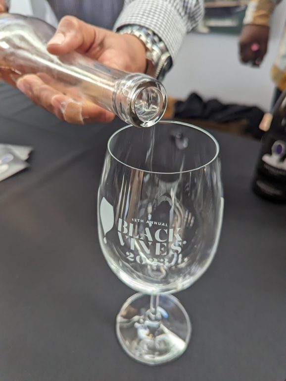 A close up of a nice wine glass with the words "Black Vines 2023" on it while someone is pouring a white wine