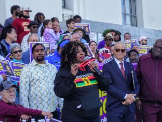 An African American woman with curly hair holding red sunglasses speaks into a mic in front of a courthouse
