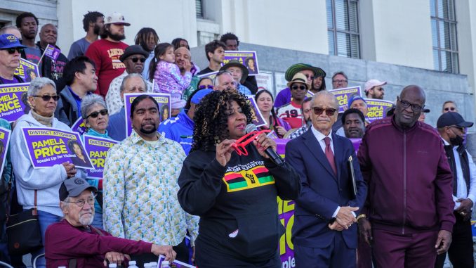 An African American woman with curly hair holding red sunglasses speaks into a mic in front of a courthouse