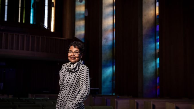An elder African American woman with short hair and a dress smiles for camera inside a church with colorful windows