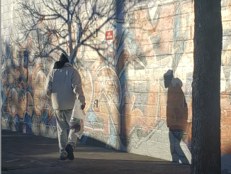 A man walks past a wall slightly hunched over while he casts a shadow against the wall