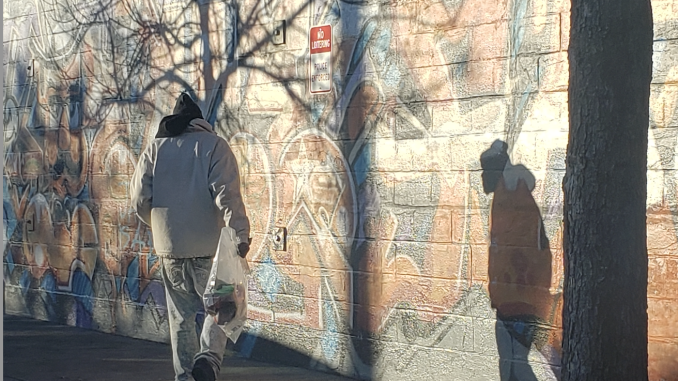 A man walks past a wall slightly hunched over while he casts a shadow against the wall