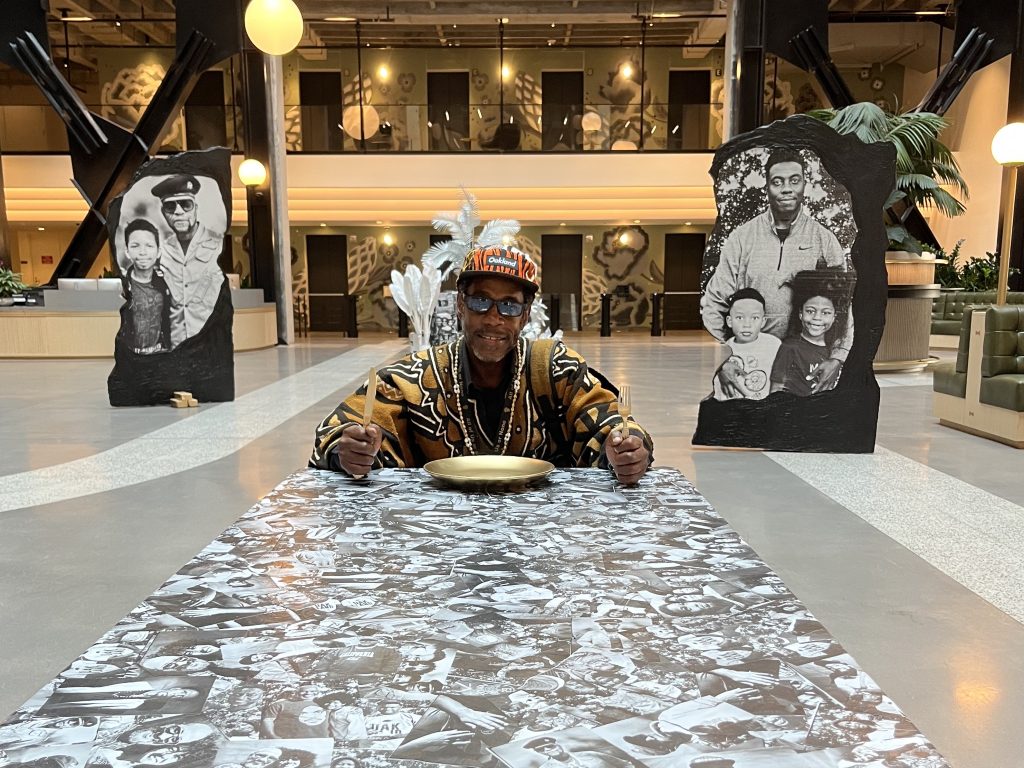 A Black man sits at the head of a table that is covered with black and white images and collages of Black fathers
