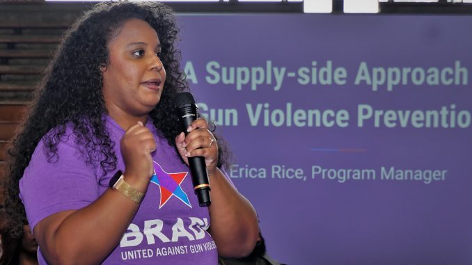 An African American woman with curly hair holding a mic speaks in front of a purple display about gun violence