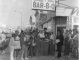 a black and white photo of a storefront that says Everett & jones Bar-B-Q and a group of Black people dressed in 70s attire pose in front of it