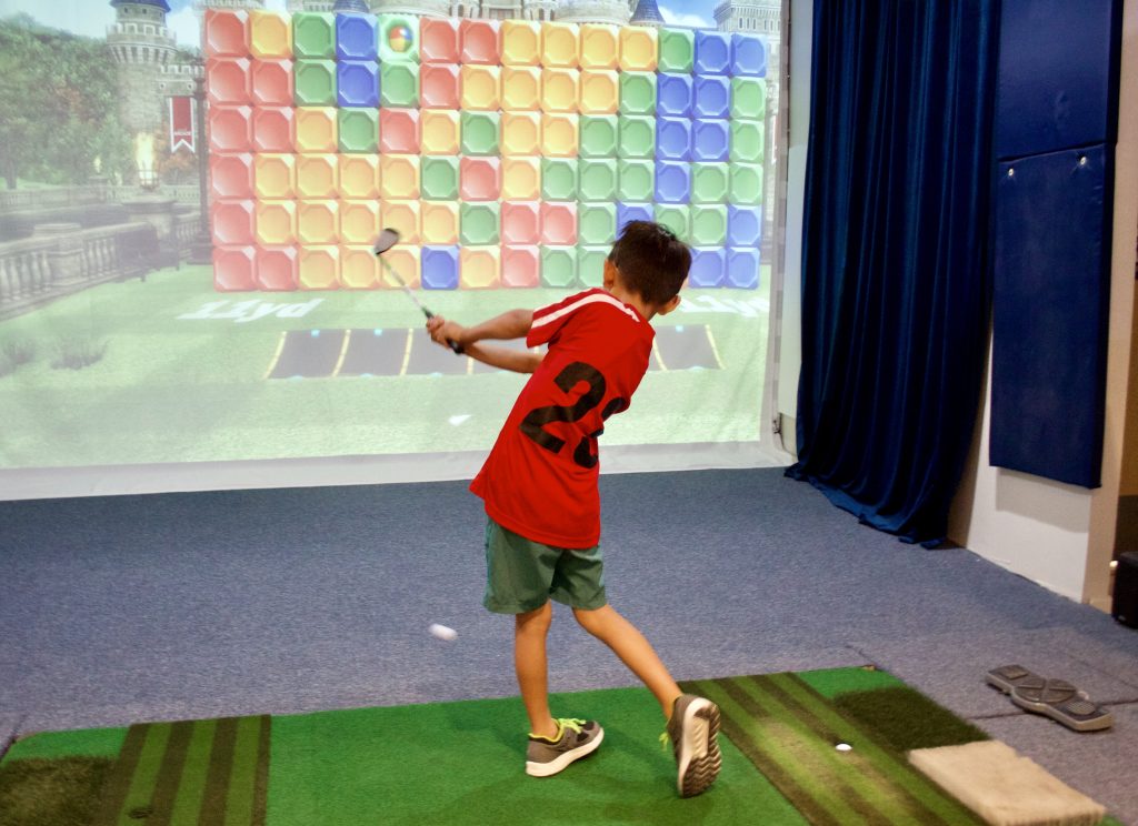 A young boy wearing a red jersey shirt swings his golf club into a simulator screen