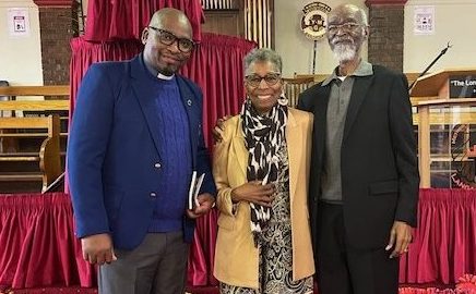 A Black pastor, and elder African American woman, and elder African American man pose for a photo inside a church with red carpet