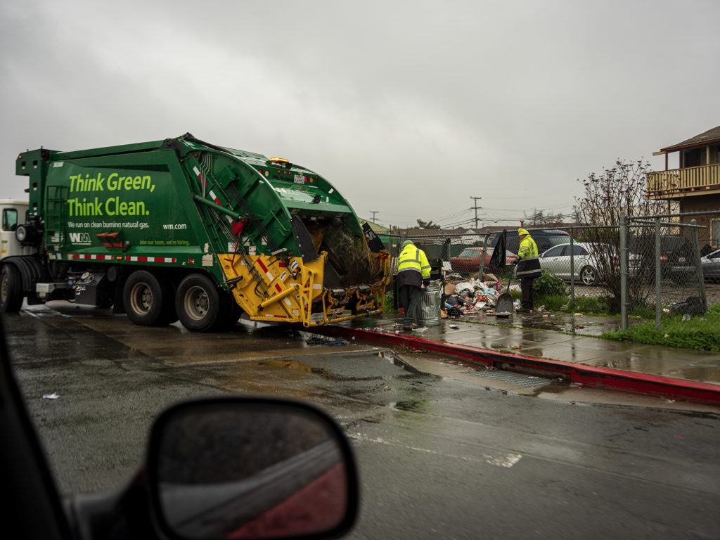 A large green garbage truck picks up trash from a sidewalk. a rearview mirror in the foreground