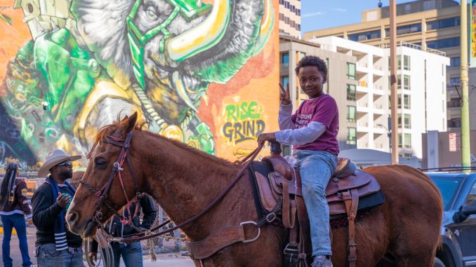 A young Black boy sits on a horse and shows a peace sign with his hand
