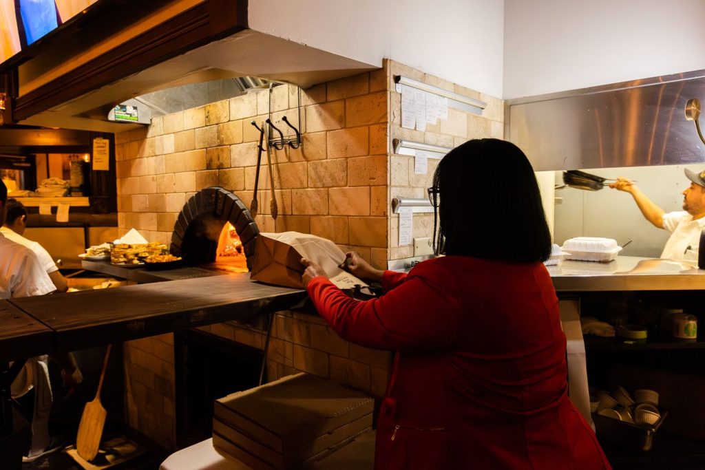 A woman with a red jacket packs up a to-go mean in front of a wood oven