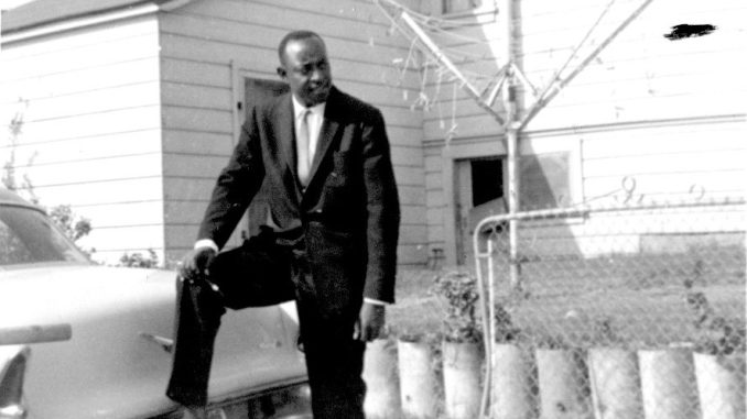 a black and white photo of a Black man in a suit in front of a house