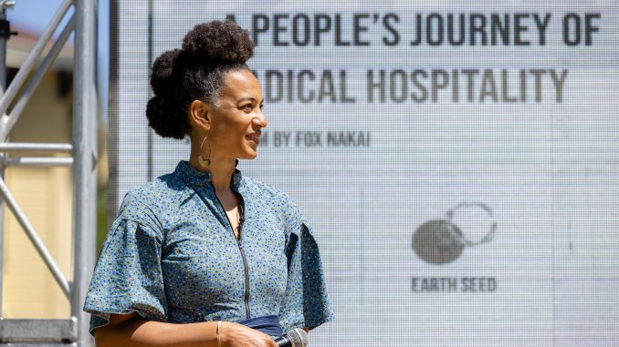 A Black woman stands on an outdoor stage with a microphone.