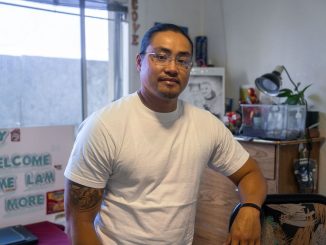 An Asian American man sits in a chair inside a room with a sign that says "no more" and a photo of a ramen packet.