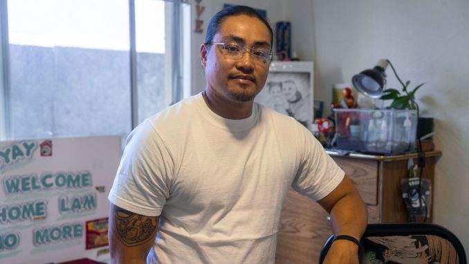 An Asian American man sits in a chair inside a room with a sign that says "no more" and a photo of a ramen packet.