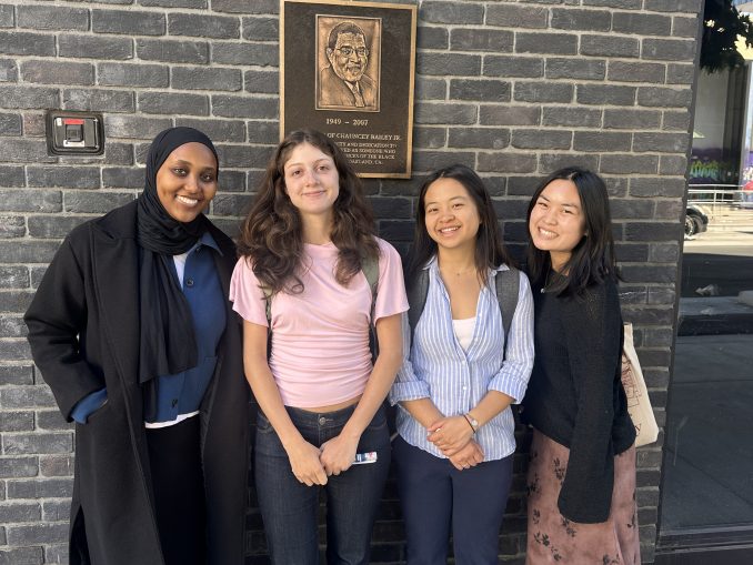 five smiling young women standing in front of plaque in downtown oakland
