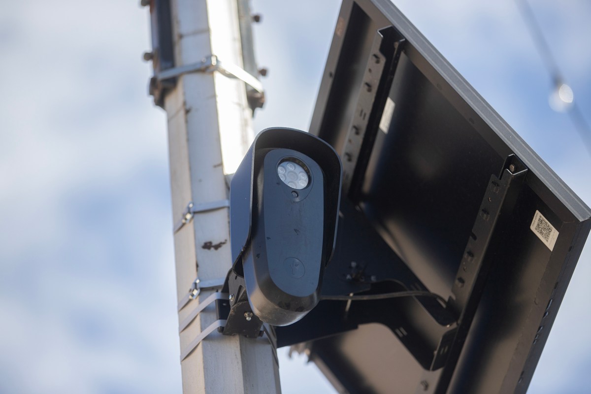 A close-up view shows a surveillance camera mounted to a metal pole beside a solar panel. The camera points outward against a bright sky, with mounting hardware and wiring visible.